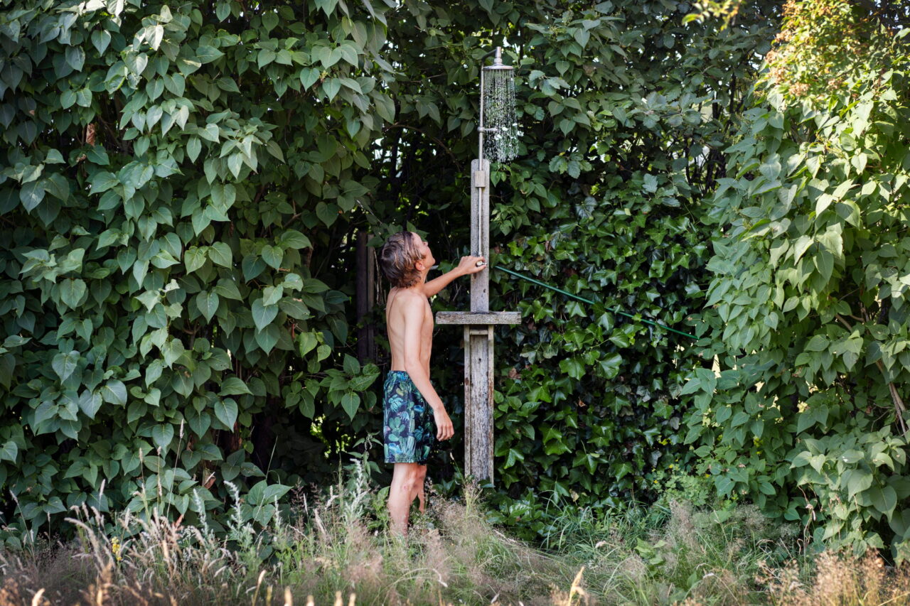 Intill en utedusch av grånat trä står en pojke och håller sin hand på kranen. Vattnet har glittrande börjat rinna.