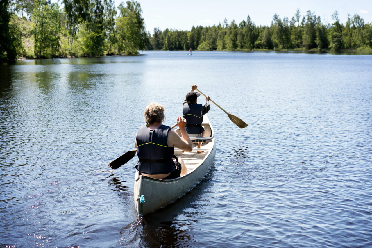 På väg bort paddlar två personer i en kanot. Klädda i flytväst och med paddlarna på var sin sida i det djupblå vattnet.