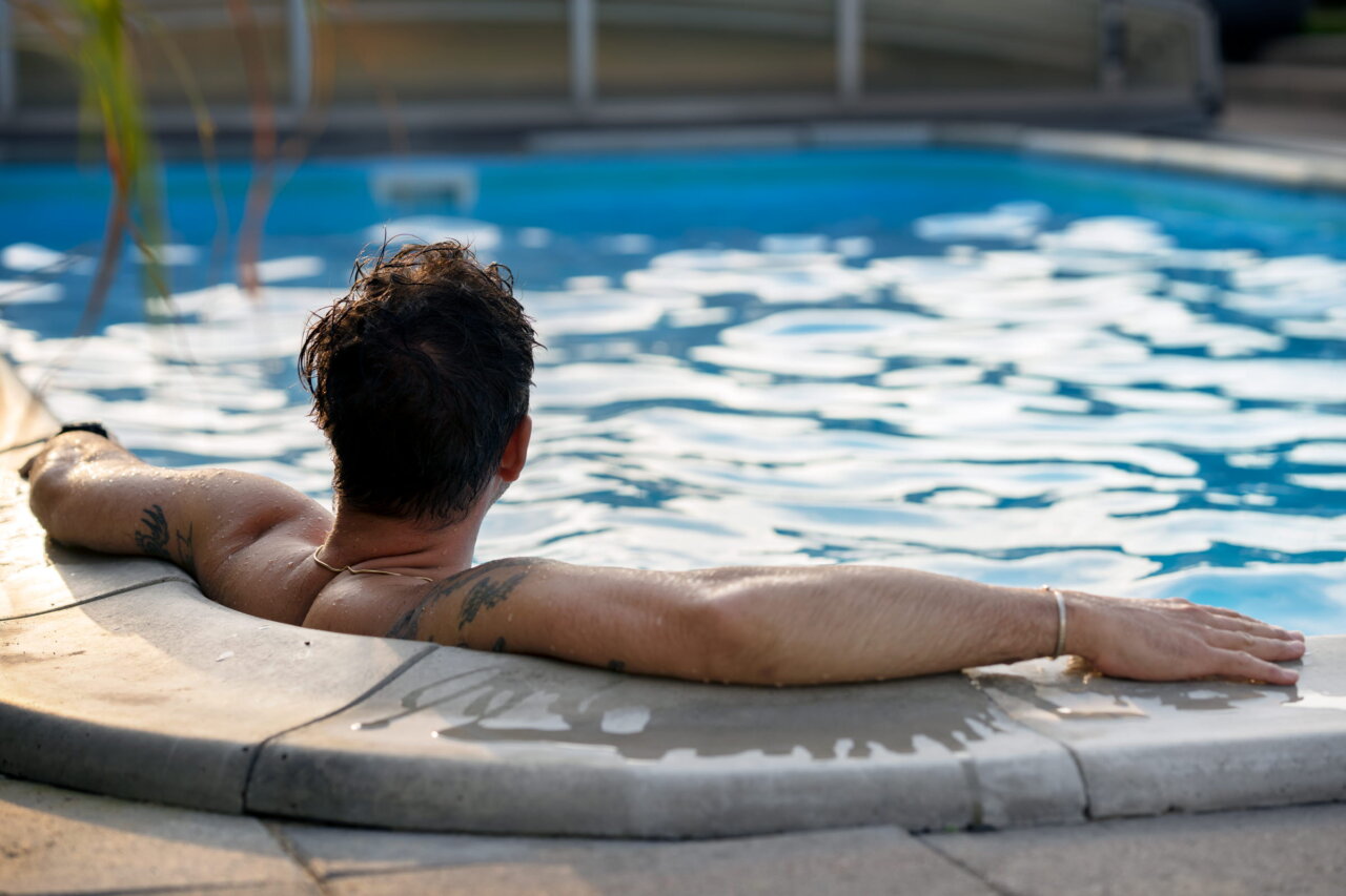 En man sitter i poolens rundade hörn. Armar och händer vilar på kanten. Håret är rufsigt och blött.